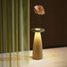 Flora cordless table lamp with LED light and flower vase design on marble surface near green curtain