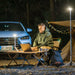 Man using laptop at night outdoors illuminated by multi-functional camping light on tripod near tent and car