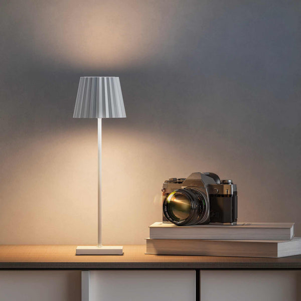 Plissé cordless table lamp with white pleated shade on wooden surface next to camera and books, modern LED rechargeable design