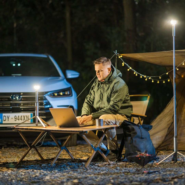 Man using laptop at night outdoors illuminated by multi-functional camping light on tripod near tent and car